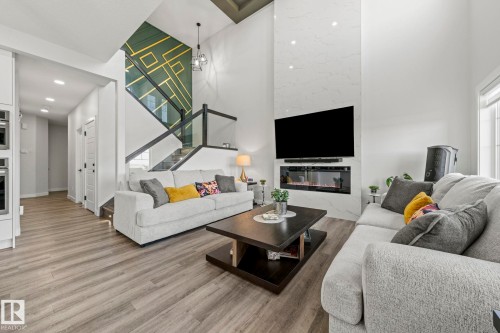 Open-concept living area featuring high ceilings, wood-finish flooring, a contemporary fireplace, and a striking accent wall with geometric detailing - 16128 31 Avenue Sw, Edmonton, AB - Indoor Photo Showing Living Room With Fireplace