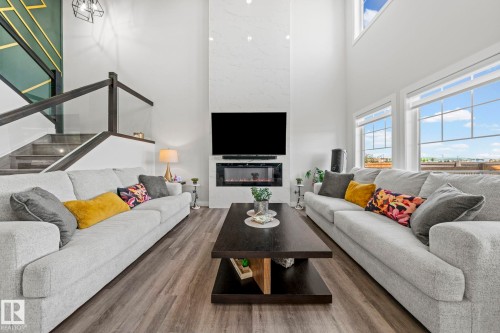 Two-story living area featuring a prominent fireplace with a light-toned surround, extensive window banks, wood-finish flooring, and a contemporary open-riser staircase with glass panels - 16128 31 Avenue Sw, Edmonton, AB - Indoor Photo Showing Living Room With Fireplace