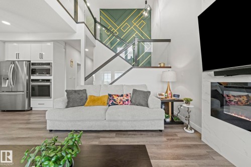 Wood-finish flooring, built-in electric fireplace with stone surround, stainless steel built-in double oven, stainless steel refrigerator, and custom accent wall with geometric design - 16128 31 Avenue Sw, Edmonton, AB - Indoor Photo Showing Living Room With Fireplace