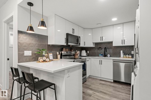 White high-gloss cabinetry, stainless steel appliances, textured dark gray backsplash, light quartz countertops, and wood-finish flooring - 16128 31 Avenue Sw, Edmonton, AB - Indoor Photo Showing Kitchen With Upgraded Kitchen