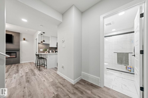 Modern kitchen with white cabinetry and pendant lighting, fireplace with tile surround, wood-finish flooring, and a bathroom with a glass shower enclosure - 16128 31 Avenue Sw, Edmonton, AB - Indoor With Fireplace