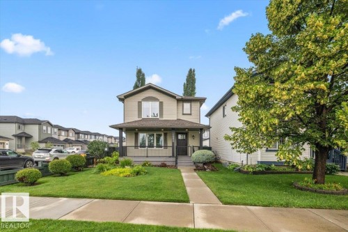 Two-story residence featuring a covered front porch with black railings, a light tan facade with dark green shutters, and a mature tree in the front yard - 1086 Grantham Drive Nw, Edmonton, AB - Outdoor With Deck Patio Veranda With Facade