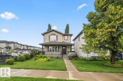 Two-story residence featuring a covered front porch with black railings, a light tan facade with dark green shutters, and a mature tree in the front yard - 