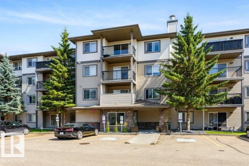 Multi-level building with individual balconies, prominent evergreen landscaping, ample exterior parking, and a stone-clad entrance - #307 1188 Hyndman Rd Nw, Edmonton, AB - Outdoor With Balcony With Facade
