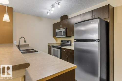 Kitchen featuring stainless steel appliances, dark wood cabinetry, a double basin sink with a high-arc faucet, and track lighting - #307 1188 Hyndman Rd Nw, Edmonton, AB - Indoor Photo Showing Kitchen With Double Sink