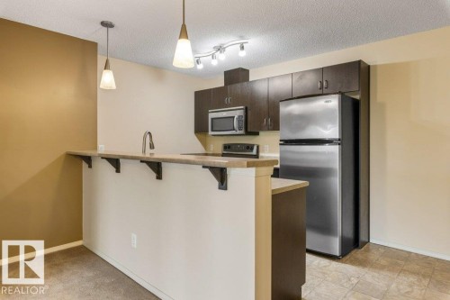 Kitchen featuring dark wood-finish cabinetry, stainless steel appliances, and a breakfast bar - #307 1188 Hyndman Rd Nw, Edmonton, AB - Indoor Photo Showing Kitchen
