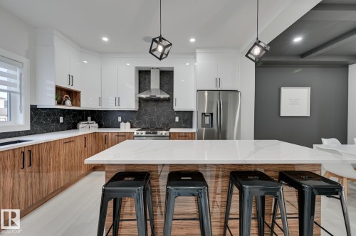 Modern kitchen featuring white and wood-finish cabinetry, a large center island with a white countertop, stainless steel appliances, and a black patterned backsplash - 51 Clichy Close, St. Albert, AB - Indoor Photo Showing Kitchen With Upgraded Kitchen