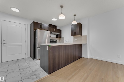 Contemporary kitchen featuring dark wood-finish cabinetry, stainless steel appliances, and a light-toned countertop - #217 667 Watt Bv Sw, Edmonton, AB - Indoor Photo Showing Kitchen