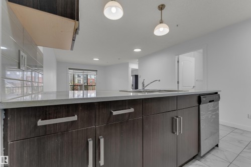 Kitchen island featuring dark wood-finish cabinetry, light-toned countertops, an undermount sink with a gooseneck faucet, a built-in stainless steel dishwasher, and two pendant light fixtures - #217 667 Watt Bv Sw, Edmonton, AB - Indoor Photo Showing Kitchen