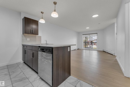 Modern kitchen featuring dark wood-finish cabinetry, light stone-finish countertops, stainless steel appliances, and pendant lighting - #217 667 Watt Bv Sw, Edmonton, AB - Indoor Photo Showing Kitchen