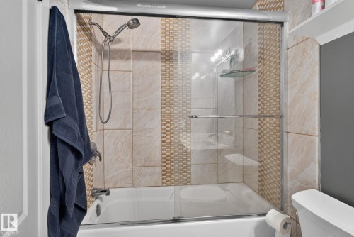 Bathtub with a sliding glass shower door and a tiled surround featuring a decorative mosaic accent - 1344 Lakewood Road West Nw, Edmonton, AB - Indoor Photo Showing Bathroom