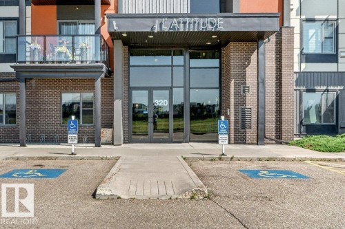 Building exterior featuring brick and paneling, a prominent glass entrance with a covered awning, and a ground-floor balcony with a glass railing - #423 320 Ambleside Li Sw, Edmonton, AB - Outdoor With Balcony