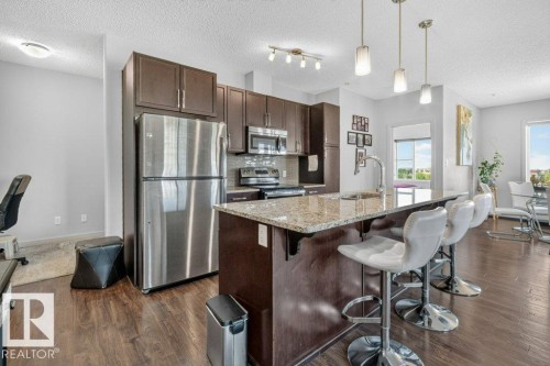 Kitchen featuring wood-finish flooring, stainless steel appliances, granite countertops, dark wood-finish cabinetry, and a tile backsplash - #423 320 Ambleside Li Sw, Edmonton, AB - Indoor Photo Showing Kitchen With Upgraded Kitchen