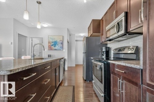 Kitchen featuring wood-finish flooring, dark wood cabinetry, granite countertops, stainless steel appliances, and a tiled backsplash - #423 320 Ambleside Li Sw, Edmonton, AB - Indoor Photo Showing Kitchen With Upgraded Kitchen