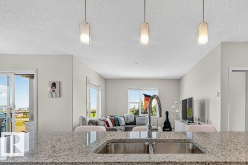 Open-concept kitchen and living area featuring pendant lighting, quartz countertop with dual undermount sinks, stainless steel appliances, and expansive windows with natural light throughout the space - #423 320 Ambleside Li Sw, Edmonton, AB - Indoor Photo Showing Kitchen With Double Sink With Upgraded Kitchen