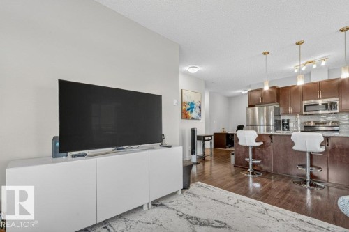 Open concept living area with wood-finish flooring, a kitchen featuring dark wood cabinetry, a tile backsplash, stainless steel appliances, and pendant lighting - #423 320 Ambleside Li Sw, Edmonton, AB - Indoor Photo Showing Kitchen