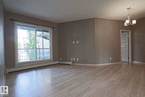 Spacious room featuring light wood-finish flooring, neutral wall paint, a sliding glass door leading to a balcony, and an overhead chandelier light fixture - #209 304 Ambleside Link Li Sw Sw, Edmonton, AB - Indoor Photo Showing Other Room