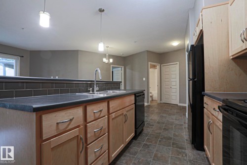 Spacious kitchen featuring a large center island with wood-finish cabinetry and drawer storage, dark tile countertops, stainless steel appliances including a range and refrigerator, subway tile backsplash, pendant lighting, and tile flooring throughout - #209 304 Ambleside Link Li Sw Sw, Edmonton, AB - Indoor Photo Showing Kitchen With Double Sink