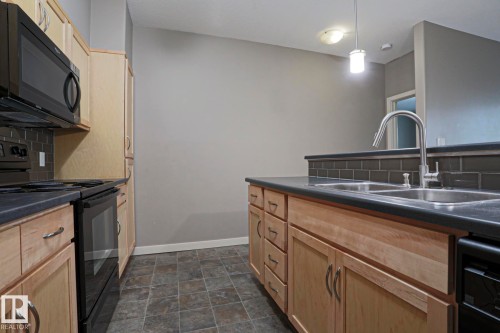 Kitchen featuring wood cabinetry, dark countertops, a subway tile backsplash, and a double basin sink with a high-arc faucet - #209 304 Ambleside Link Li Sw Sw, Edmonton, AB - Indoor Photo Showing Kitchen With Double Sink