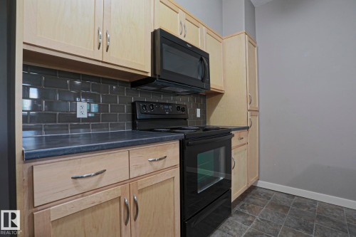 Light wood-finish cabinetry with silver-tone hardware, dark subway tile backsplash, black countertop, black range, and black microwave - #209 304 Ambleside Link Li Sw Sw, Edmonton, AB - Indoor Photo Showing Kitchen