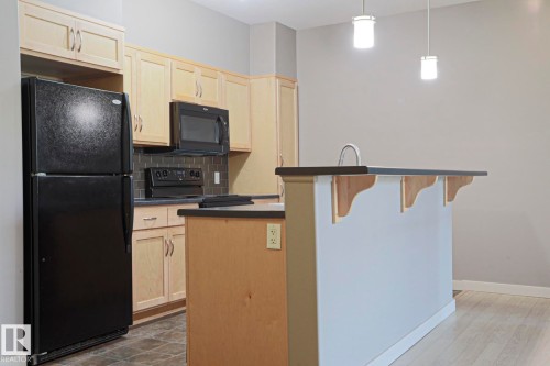 Kitchen featuring light wood-finish cabinetry, black appliances, a dark tile backsplash, and an island with an integrated sink and pendant lighting - #209 304 Ambleside Link Li Sw Sw, Edmonton, AB - Indoor Photo Showing Kitchen