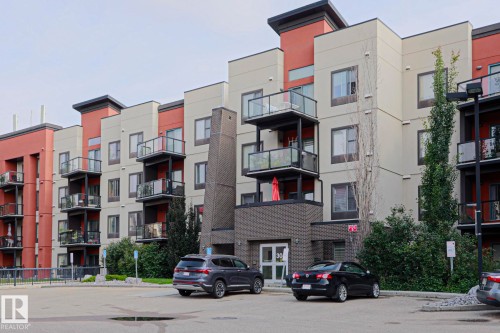 Multi-story building featuring a red and beige facade, multiple balconies with glass railings, and a brick-finish main entrance - #209 304 Ambleside Link Li Sw Sw, Edmonton, AB - Outdoor With Balcony With Facade