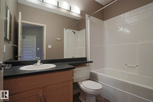 Integrated vanity with a dark countertop and undermount sink, wood-finish cabinetry, and a full-width mirror - #209 304 Ambleside Link Li Sw Sw, Edmonton, AB - Indoor Photo Showing Bathroom