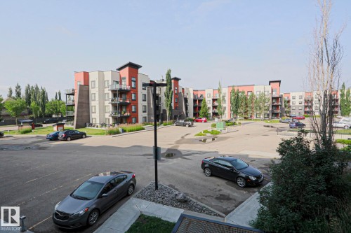 Contemporary multi-story building featuring balconies with metal railings, a paved parking area, and landscaping with mature trees and shrubs - #209 304 Ambleside Link Li Sw Sw, Edmonton, AB - Outdoor With Balcony