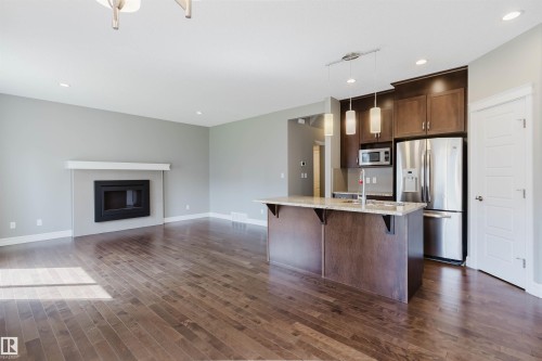 Open-concept living area featuring wood-finish flooring, a built-in fireplace with white mantel, a kitchen island with stone countertop, dark wood cabinetry, and stainless steel appliances - 15839 10 Avenue Sw, Edmonton, AB - Indoor