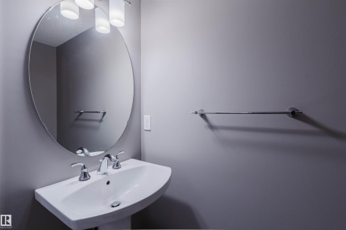 Pedestal sink with chrome fixtures, large oval mirror, and wall-mounted light fixture - 15839 10 Avenue Sw, Edmonton, AB - Indoor Photo Showing Bathroom