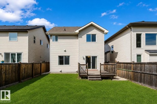 Expansive green lawn with a wooden privacy fence, featuring a two-story exterior with light-colored siding, multiple windows, and a ground-level wooden deck with steps - 15839 10 Avenue Sw, Edmonton, AB - Outdoor With Exterior