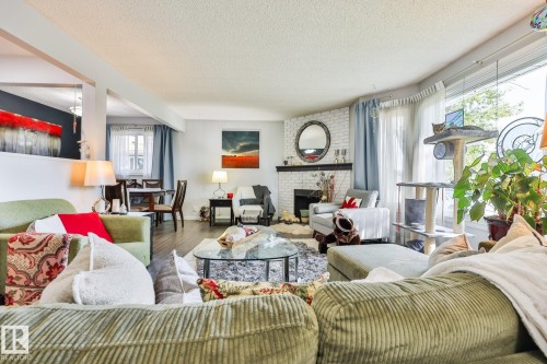 Open-concept living area with wood-finish flooring, a white brick fireplace with a dark mantel, and large windows - 8932 187 Street Nw, Edmonton, AB - Indoor Photo Showing Living Room With Fireplace