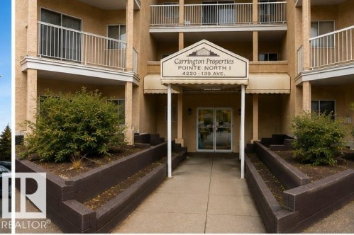 Carrington Properties North I entrance featuring a welcoming covered portico with white trim detailing, dual planters with established landscaping flanking the entry, and a multi-story brick facade with metal railings on upper-level balconies - #306 4220 139 Av Nw, Edmonton, AB - Outdoor