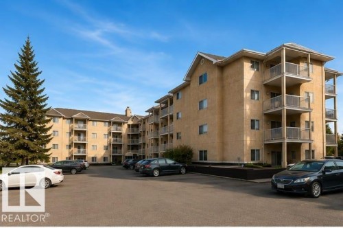 Multi-unit residential complex featuring stucco exterior with balconies, ample parking area, mature evergreen landscaping, and well-proportioned architectural design across multiple stories - #306 4220 139 Av Nw, Edmonton, AB - Outdoor With Facade