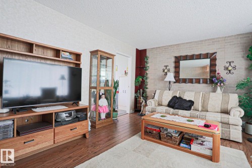 Living area featuring wood-finish flooring, a textured white ceiling, and an accent wall with a light-colored brick pattern - 5203 45 Street, Beaumont, AB - Indoor Photo Showing Living Room