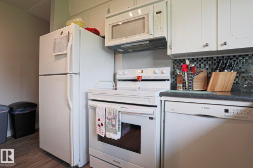 Kitchen featuring white cabinetry, dark countertops, a mosaic tile backsplash, wood-finish flooring, and a suite of white appliances including a refrigerator, range, microwave, and dishwasher - 5203 45 Street, Beaumont, AB - Indoor Photo Showing Kitchen