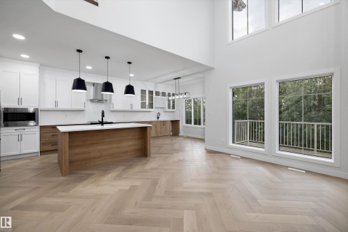 Herringbone wood-finish flooring, two-story living space with multiple windows, kitchen with white upper cabinetry and wood-finish lower cabinetry, kitchen island with built-in sink, and pendant lighting - 971 Elderberry Landing Nw, Edmonton, AB - Indoor Photo Showing Other Room