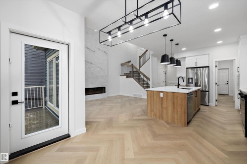 Herringbone wood-finish flooring, a kitchen island with a white countertop, stainless steel appliances, a white marble-patterned fireplace surround, and a staircase with wood treads and metal balusters - 971 Elderberry Landing Nw, Edmonton, AB - Indoor Photo Showing Kitchen