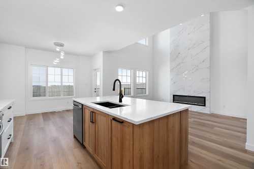 Kitchen island with a built-in sink and dishwasher, white countertops, wood-finish flooring, white walls, and a tall fireplace with a linear insert - 1763 Crossbill Loop Nw, Edmonton, AB - Indoor Photo Showing Kitchen With Double Sink
