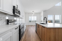 Kitchen featuring white shaker cabinetry, stainless steel appliances, marble-look backsplash, wood-finish flooring, and a central island with a contrasting wood-finish base and integrated sink - 