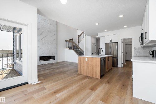 Spacious open-concept interior featuring light wood-finish flooring, a contemporary linear fireplace with a striking floor-to-ceiling tile surround, and a kitchen island with a wood-finish base and light-toned countertop - 1763 Crossbill Loop Nw, Edmonton, AB - Indoor Photo Showing Kitchen With Upgraded Kitchen