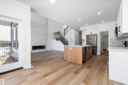 Spacious open-concept interior featuring light wood-finish flooring, a contemporary linear fireplace with a striking floor-to-ceiling tile surround, and a kitchen island with a wood-finish base and light-toned countertop - 