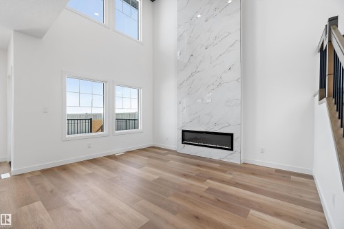 Double-height ceiling, wood-finish flooring, two large windows, two transom windows, and a linear electric fireplace with a marble-finish surround - 1763 Crossbill Loop Nw, Edmonton, AB - Indoor With Fireplace