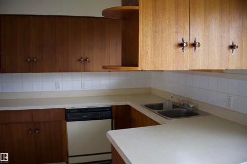 Kitchen featuring wood-finish cabinetry, light-toned countertops, a stainless steel double sink, white tiled backsplash, and a built-in dishwasher - 4946 47 Street, Barrhead, AB - Indoor Photo Showing Kitchen With Double Sink