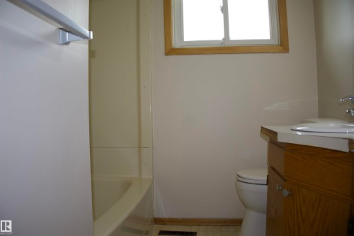 Compact bathroom featuring a wood-finish vanity with integrated sink, ceramic tile walls, a separate toilet, and a window providing natural light - 4946 47 Street, Barrhead, AB - Indoor Photo Showing Bathroom