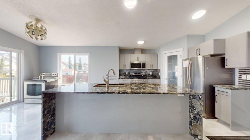 Kitchen featuring a large central island with a waterfall countertop, stainless steel appliances, light gray cabinetry, and a full-height tile backsplash - 7848 7 Avenue Sw, Edmonton, AB - Indoor Photo Showing Kitchen