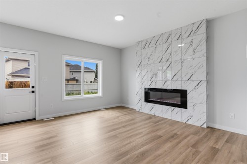Spacious room featuring light wood-finish flooring, a contemporary electric fireplace with a tiled surround, a white window, and a white exterior door - 8815 181 Avenue Nw, Edmonton, AB - Indoor Photo Showing Living Room With Fireplace