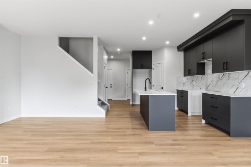 Open-concept living space featuring wood-finish flooring, a contemporary kitchen island with a built-in sink, dark cabinetry, light-toned countertops, and a white tile backsplash - 8815 181 Avenue Nw, Edmonton, AB - Indoor Photo Showing Kitchen With Upgraded Kitchen