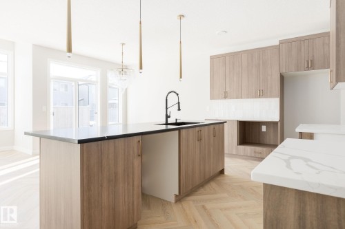 Kitchen featuring wood-finish cabinetry, white tile backsplash, a central island with a black countertop and integrated sink, and herringbone wood-finish flooring - 24 Clichy Close, St. Albert, AB - Indoor Photo Showing Kitchen