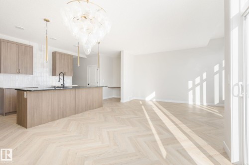 Spacious open-concept area featuring wood-finish herringbone flooring, a large kitchen island with dark countertops, light wood-finish cabinetry, and a decorative ceiling fixture with three pendant lights - 24 Clichy Close, St. Albert, AB - Indoor Photo Showing Kitchen